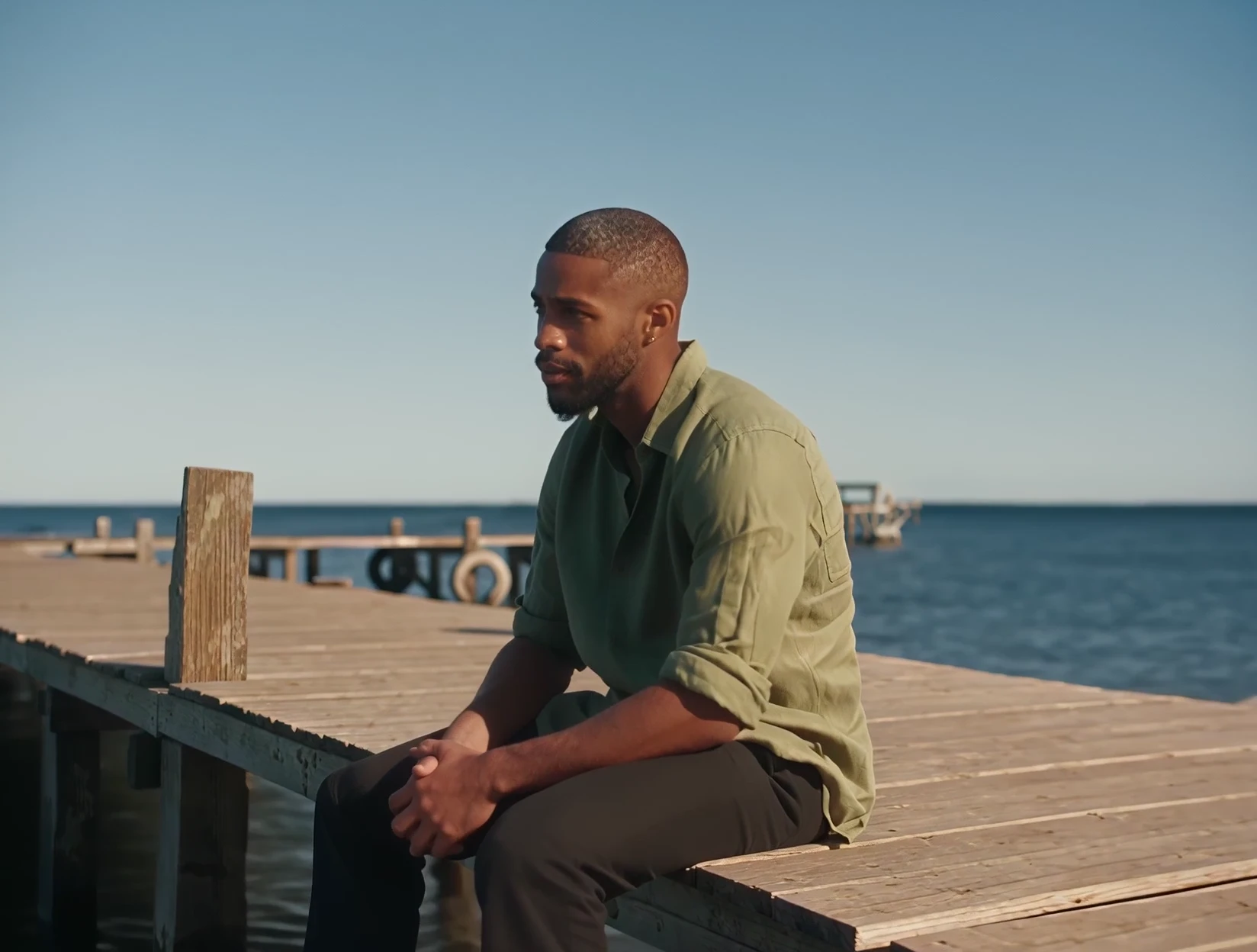 A man in a sage green linen shirt waits alone on a sunlit pier