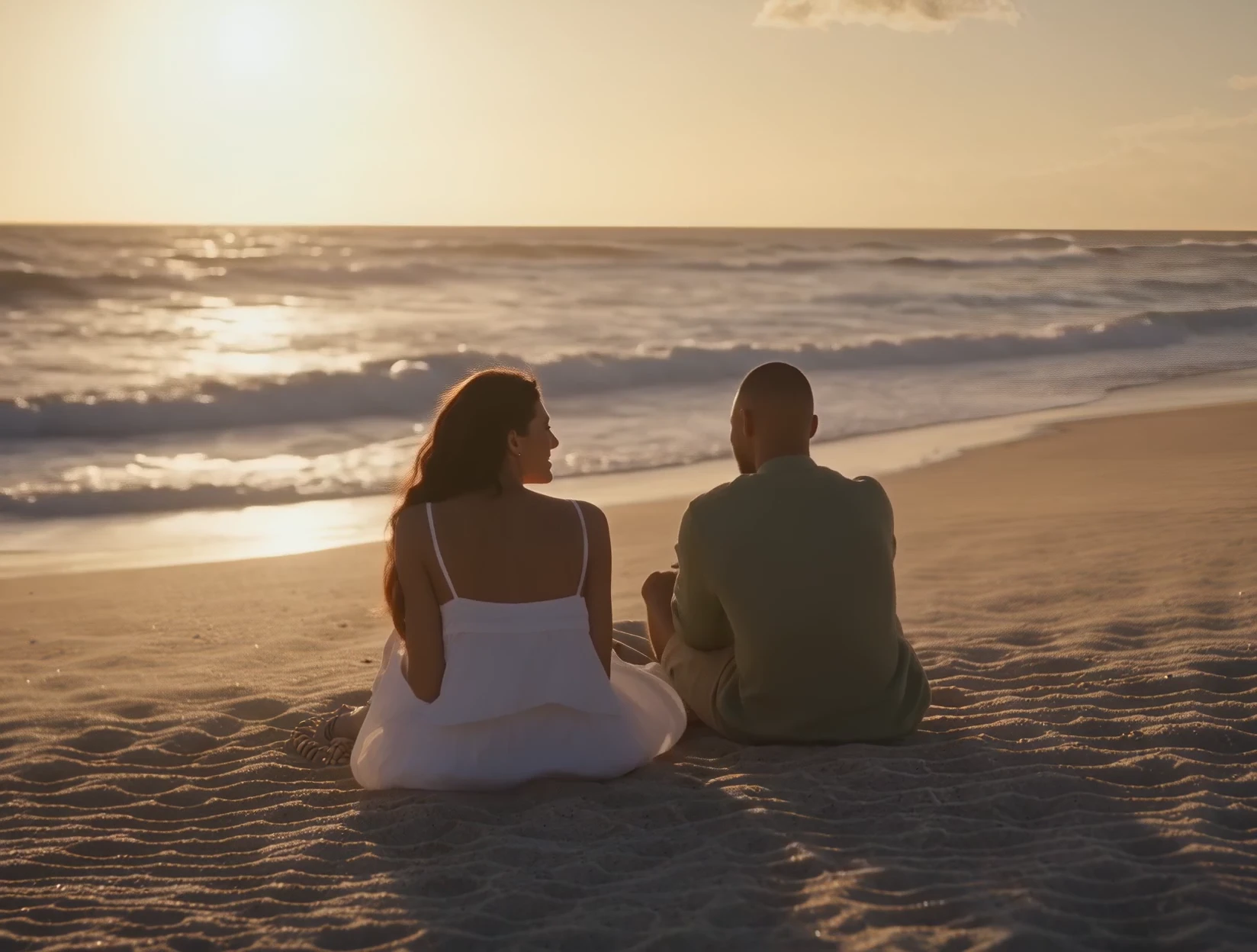 The two of them sit on the beach at sunset, seen from behind